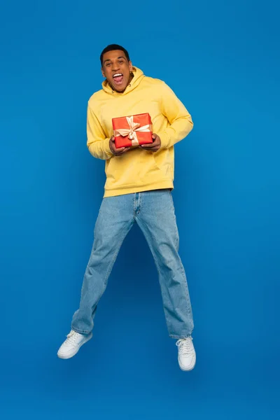 Hombre afroamericano excitado levitando con caja de regalo roja sobre fondo azul - foto de stock