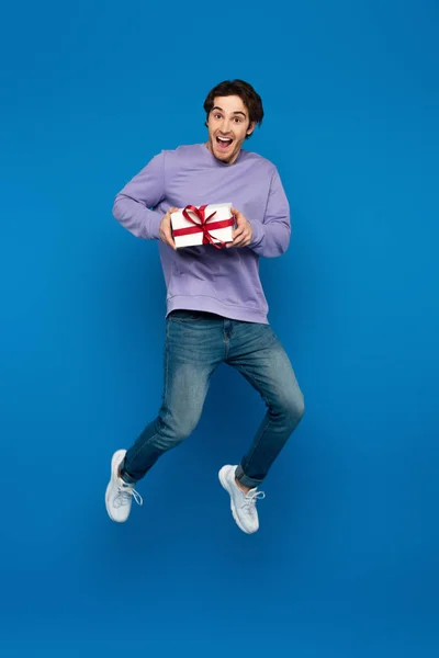 Excited young man levitating with gift box on blue background — Stock Photo