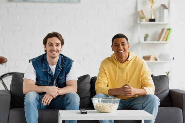Sonrientes amigos interracial sentados en el sofá, viendo la televisión y comiendo palomitas de maíz en casa - foto de stock