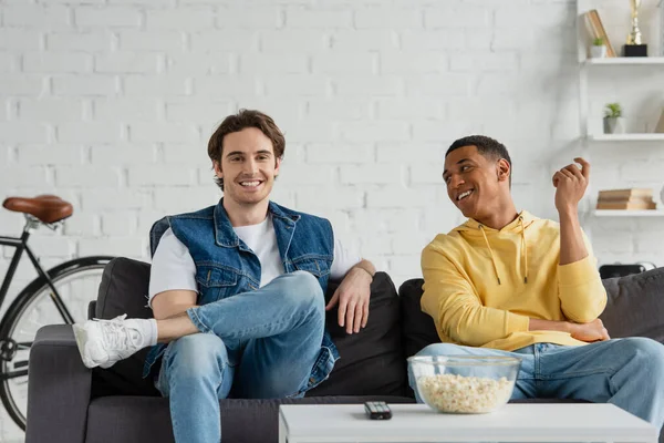 Amigos interracial feliz sentado en el sofá, viendo la televisión y comer palomitas de maíz en casa - foto de stock