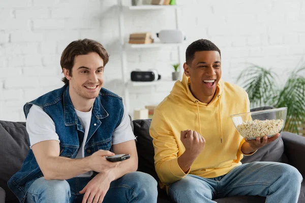 Молодые межрасовые друзья сидят на диване, смотрят телевизор и едят попкорн дома — стоковое фото