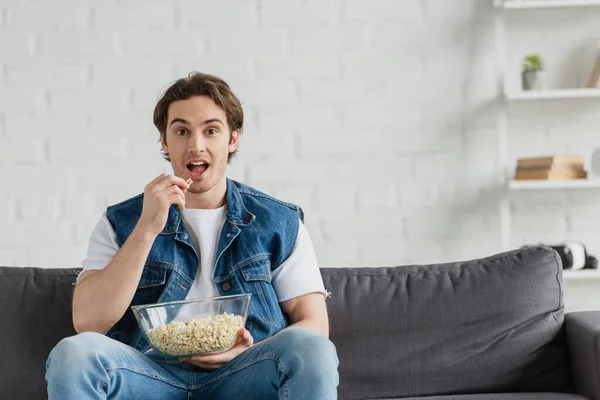 Joven sentado en el sofá, viendo la televisión y comiendo palomitas de maíz en casa - foto de stock