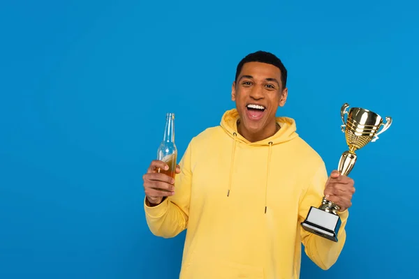 Smiling african american man holding trophy cup and beer bottle in hands isolated on blue — Stock Photo