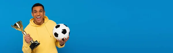 Positive african american man holding football and cup in hands isolated on blue, banner — Stock Photo