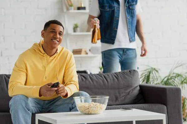 Sorridente afro-americano homem segurando tv controlador remoto e compartilhar cerveja com amigo na sala de estar moderna — Fotografia de Stock