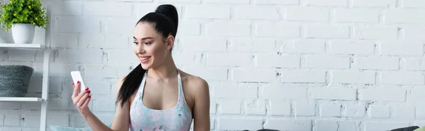 Mujer sonriente en ropa deportiva usando el teléfono inteligente en casa, bandera - foto de stock