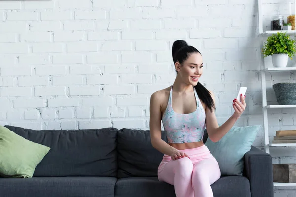 Femme sportive souriant tout en étant assis sur le canapé et en utilisant un smartphone — Photo de stock