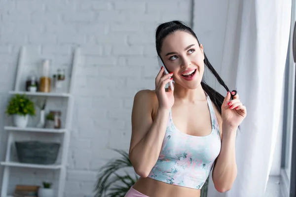 Mujer alegre y coqueta en sujetador deportivo tocando el pelo mientras habla en el teléfono móvil en casa - foto de stock