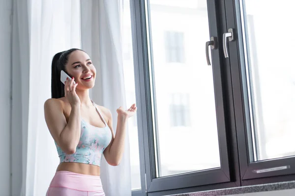 Mulher esportiva sorrindo ao falar no celular perto da janela em casa — Fotografia de Stock