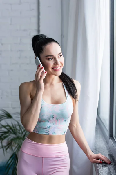Smiling woman in sports bra standing near window and talking on smartphone — Stock Photo