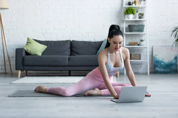 Brünette Frau in Sportbekleidung schaut auf Laptop, während sie auf Yogamatte sitzt — Stock Photo