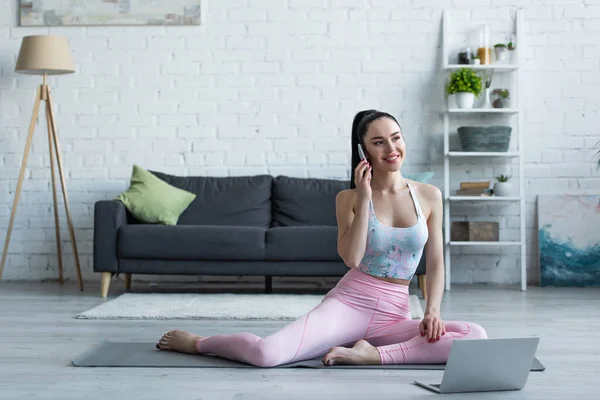 Mujer joven y feliz hablando en el teléfono móvil en la esterilla de yoga cerca del ordenador portátil - foto de stock