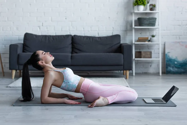 Вид збоку жінки, що практикує позу героя на килимку йоги біля ноутбука — стокове фото