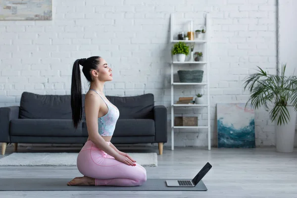 Seitenansicht der brünetten Frau in sitzender Heldenpose in der Nähe von Laptop zu Hause — Stock Photo