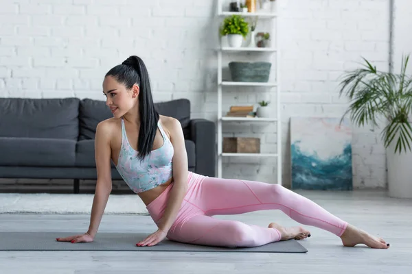 Bonita, mujer descalza sonriendo mientras está sentada en la esterilla de yoga en casa - foto de stock