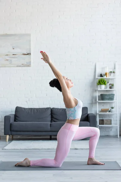 Vista lateral de la joven morena practicando media luna se abalanzan sobre la rodilla posan sobre esterilla de yoga - foto de stock