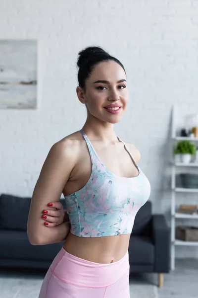 Jolie femme en soutien-gorge de sport souriant à la caméra tout en se tenant avec les mains derrière le dos — Photo de stock