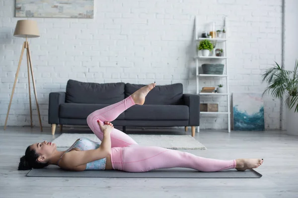 Vista laterale della donna in abbigliamento sportivo sdraiata nel vento rimuovere posa su tappetino yoga — Foto stock