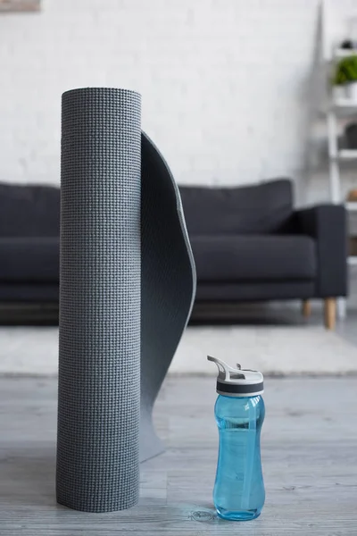 Bottiglia sportiva con acqua vicino al tappeto fitness arrotolato su sfondo sfocato — Foto stock