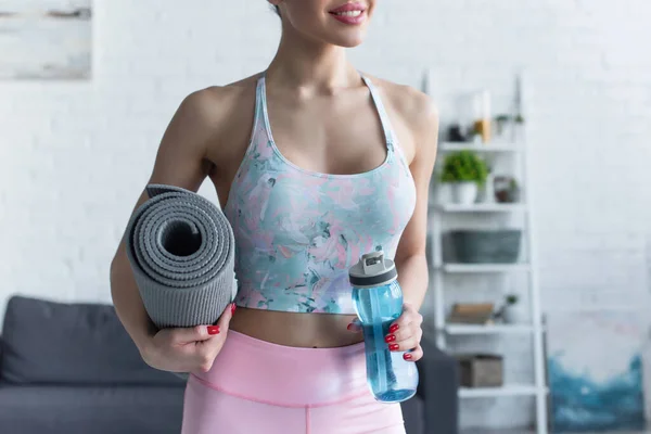 Visão parcial da mulher sorridente segurando tapete de fitness e garrafa de esportes em casa — Fotografia de Stock