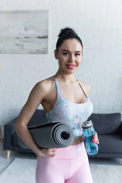 Happy woman with sports bottle and fitness mat smiling at camera — Stock Photo