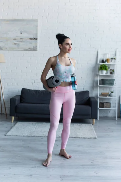 Спортивная босая женщина, стоящая с ковриком для йоги и спортивной бутылкой дома — стоковое фото