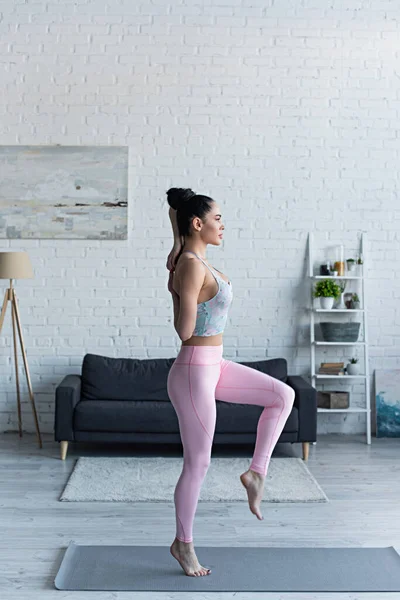 Vista lateral de la joven mujer de pie sobre una pierna con las manos detrás de la espalda mientras practica yoga en casa - foto de stock