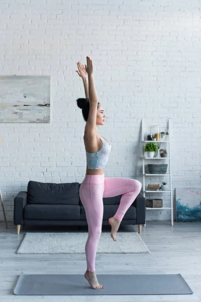 Seitenansicht einer brünetten Frau, die mit erhobenen Händen auf einem Bein steht, während sie Yoga praktiziert — Stockfoto