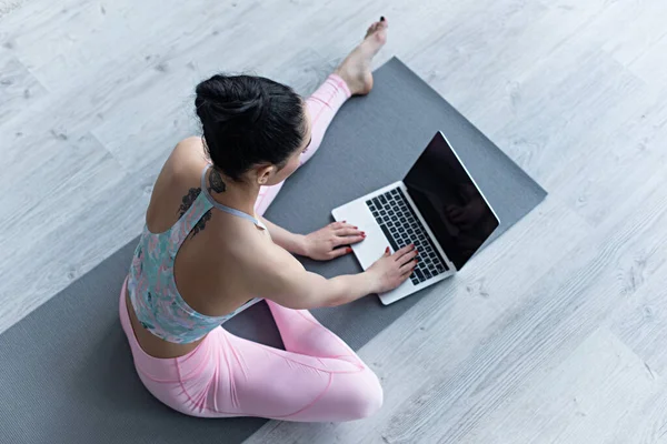 Vue aérienne de la femme en vêtements de sport en utilisant un ordinateur portable tout en étant assis sur un tapis de yoga — Photo de stock