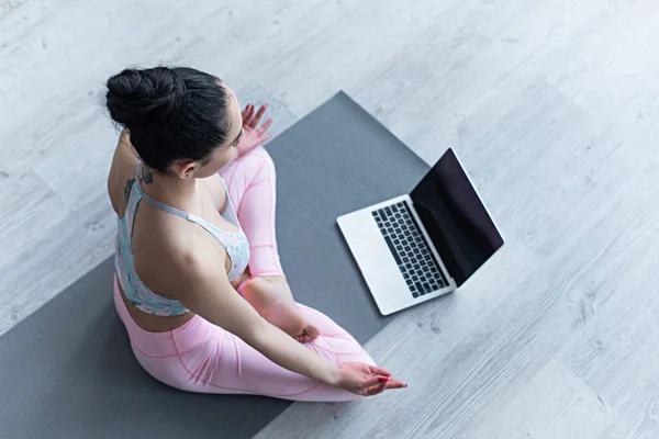 Vista aérea de la mujer meditando en la pose de loto en la esterilla de yoga cerca de la computadora portátil - foto de stock