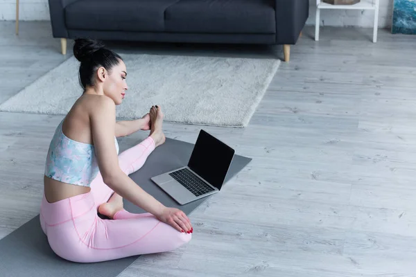 Junge Frau schaut auf Laptop, während sie sich zu Hause auf Yogamatte dehnt — Stock Photo
