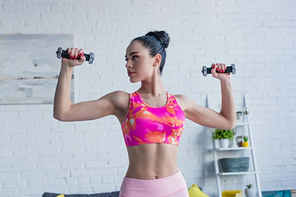 Morena mujer en sujetador deportivo mirando hacia otro lado mientras entrena con pesas en casa — Stock Photo