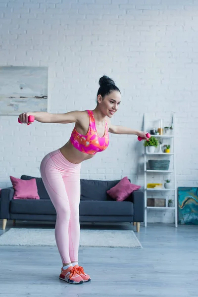 Sonriente mujer en sujetador deportivo y polainas haciendo ejercicio con mancuernas en casa — Stock Photo