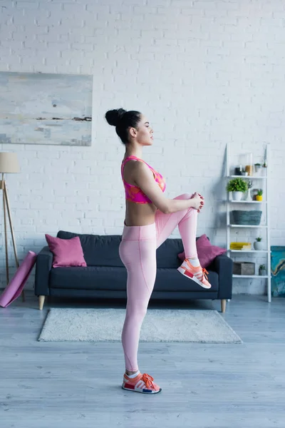 Vista lateral de una joven deportista haciendo ejercicio de rodilla a pecho posando en casa — Stock Photo