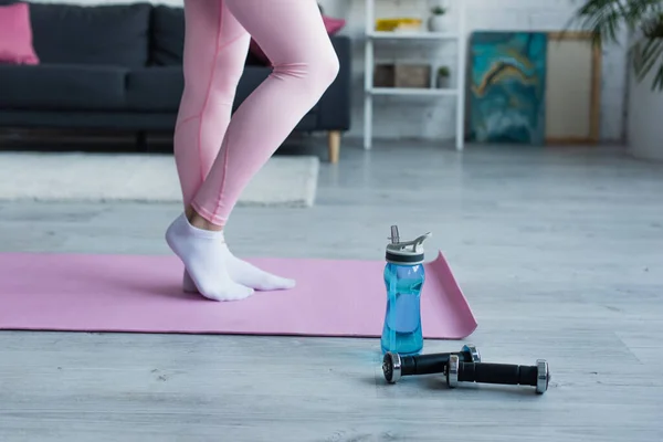 Частичный вид женщины в леггинсах и носках на фитнес-коврик возле спортивной бутылки и гантели — стоковое фото
