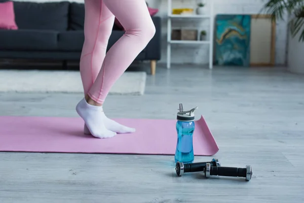 Vista parcial de la mujer en leggings y calcetines en la estera de fitness cerca de la botella de deportes y pesas - foto de stock