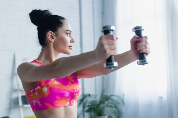 Pretty woman in sports bra looking away while training with dumbbells at home — Stock Photo