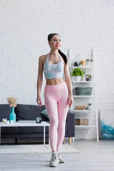 Smiling woman in sports bra and leggings looking away at home — Stock Photo
