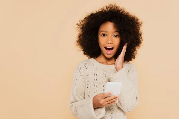 Excited african american preteen girl with hand near face holding smartphone isolated on beige — Stock Photo