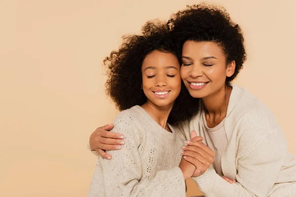 Afroamericana preadolescente hija tiernamente cogida de la mano y abrazando con la madre con los ojos cerrados sobre fondo beige - foto de stock