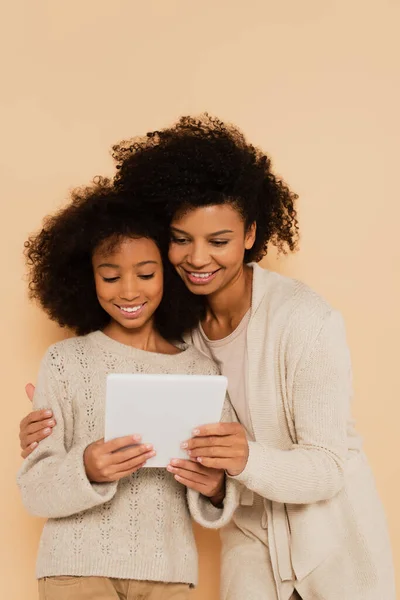 Madre afroamericana sosteniendo hija preadolescente con tableta en manos aisladas en beige - foto de stock