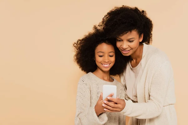 Afro-americano adulto mãe abraçando sorridente preteen filha e segurando smartphone juntos isolado no bege — Fotografia de Stock