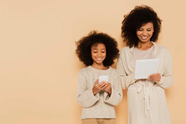 Hija preadolescente afroamericana sonriente y madre adulta sosteniendo el teléfono celular y la tableta en manos aisladas en beige — Stock Photo