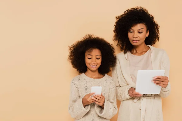 Sorrindo afro-americano preteen filha e adulto mãe segurando celular e tablet em mãos isoladas no bege — Fotografia de Stock