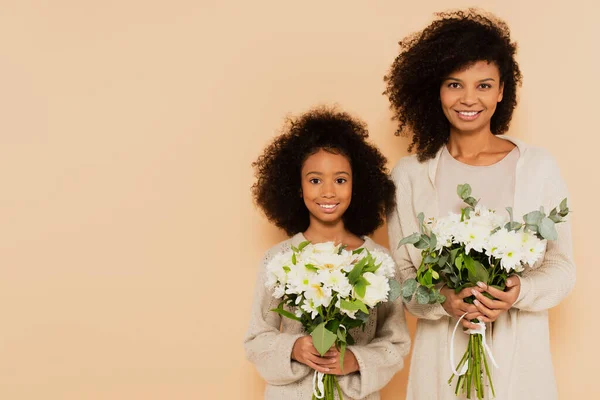 African american preteen daughter and adult mother holding bouquets of daisies isolated on beige — Stock Photo