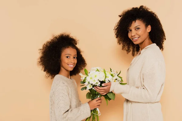 Happy african american preteen daughter and adult mother holding bouquet of daisies isolated on beige — Stock Photo