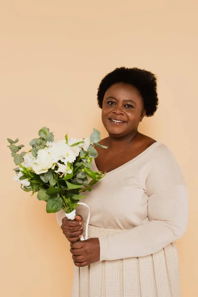 Sonriente mujer afroamericana de mediana edad en ropa pastel con ramo de flores aisladas en beige - foto de stock