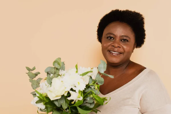 Sonriente mujer afroamericana de mediana edad con ramos de flores aisladas en beige - foto de stock