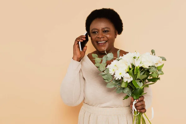 Африканская женщина среднего возраста, говорящая на смартфоне и держа букет цветов в руке на бежевом фоне — стоковое фото