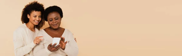 Afrikanisch-amerikanische erwachsene Tochter zeigt mit dem Finger auf Smartphone in den Händen einer Mutter mittleren Alters auf beigem Hintergrund, Banner — Stockfoto